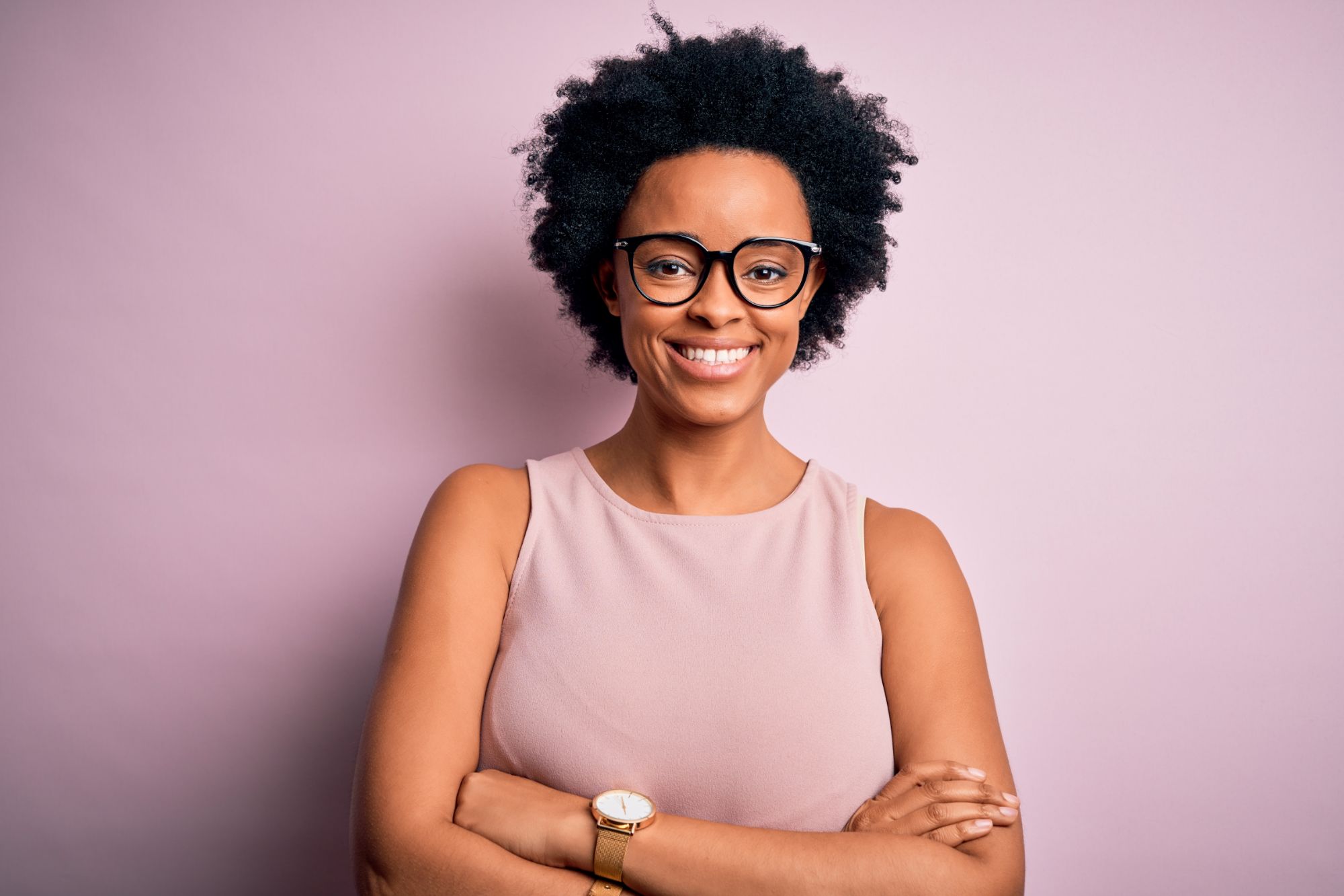 Smiling woman with glasses and natural curly hair, wearing a light pink top, against a pastel pink background, representing confidence and approachability in orthodontic care context.