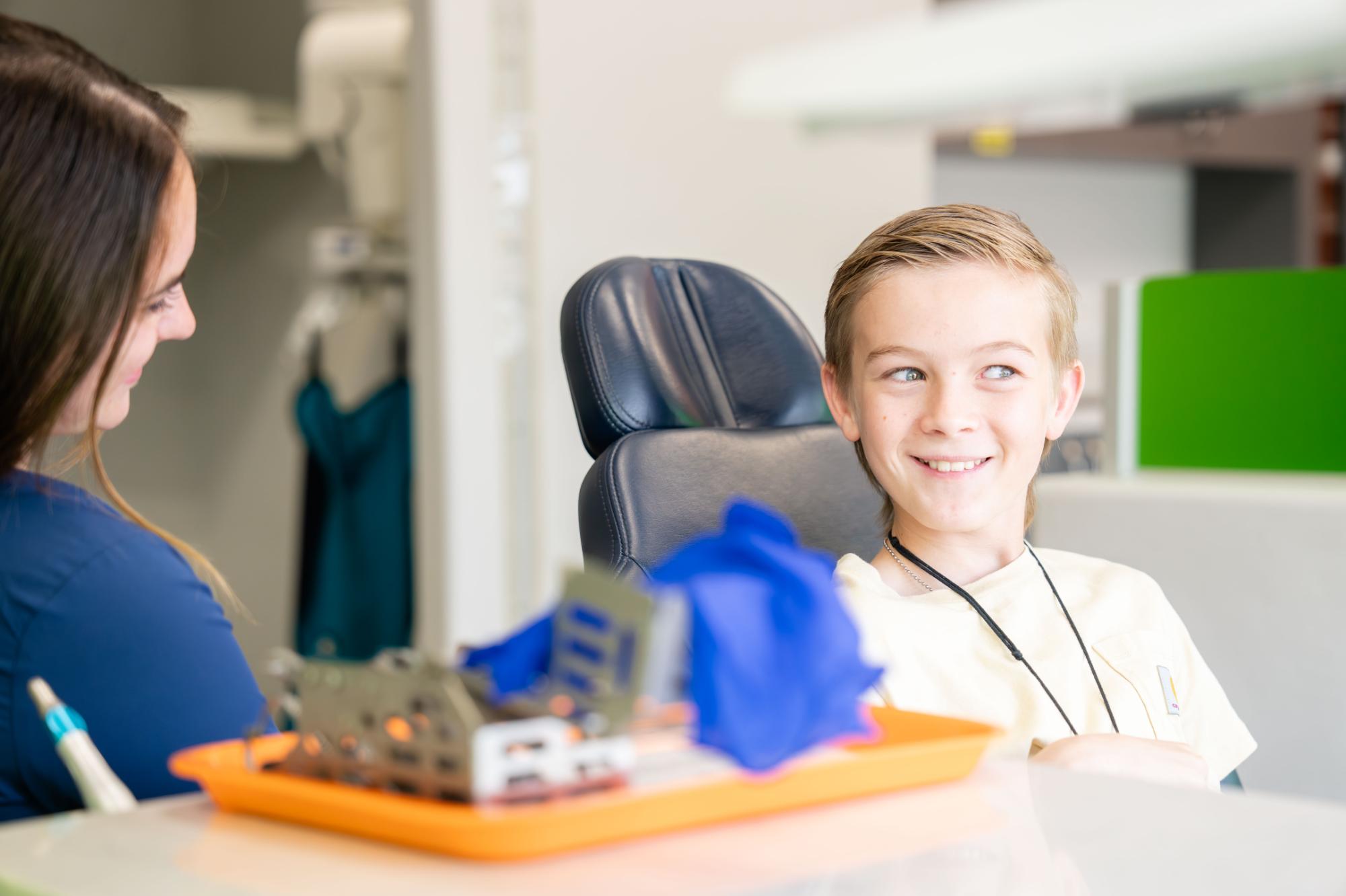Child smiling in orthodontic chair, interacting with staff member, surrounded by dental tools and equipment, reflecting a comfortable and welcoming orthodontic environment.