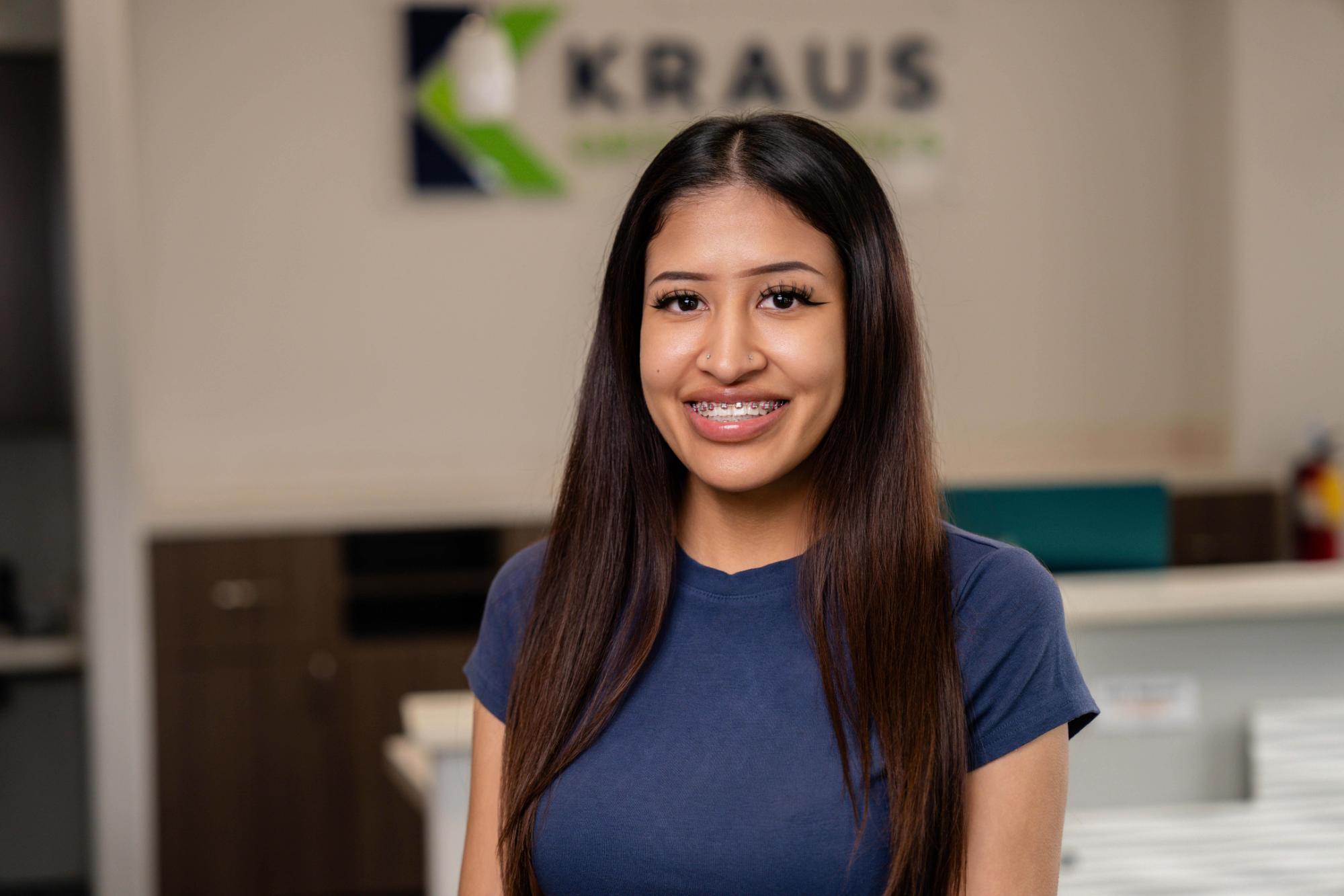 Smiling young woman with braces in a dental office, featuring Kraus Orthodontics branding in the background, showcasing patient satisfaction and orthodontic care.